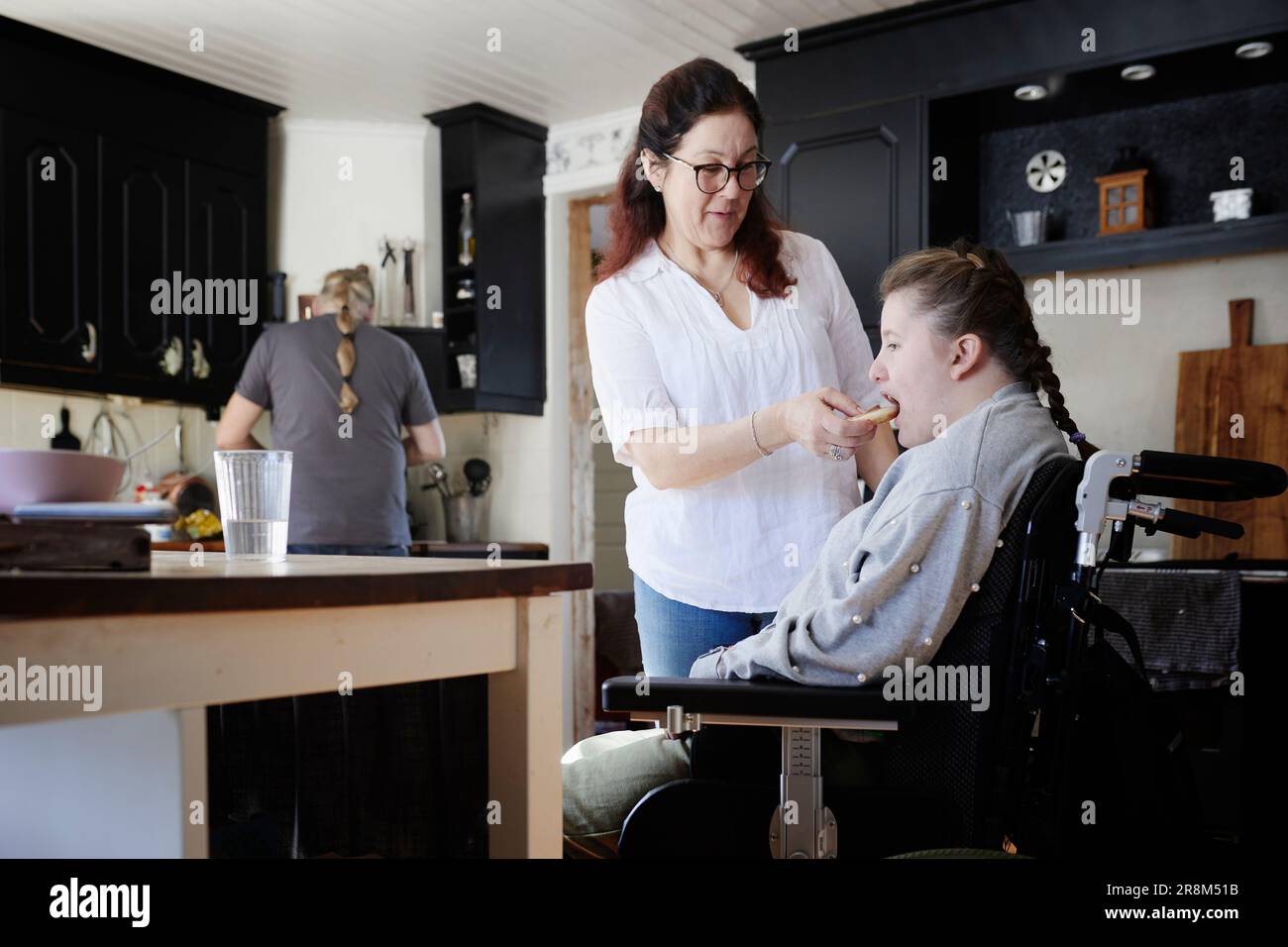 Mother feeding disabled teenage daughter in wheelchair in kitchen Stock Photo