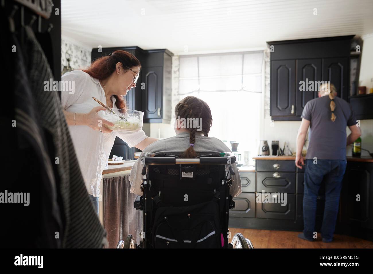 Parents with disabled teenage daughter in wheelchair in kitchen, mother feeding daughter in wheelchair while father preparing food Stock Photo