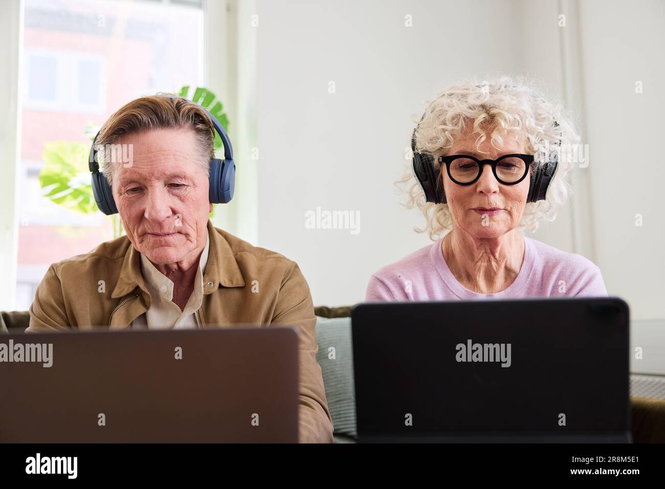 Senior man and woman sitting in living room and using laptop and digital tablet to edit podcast Stock Photo
