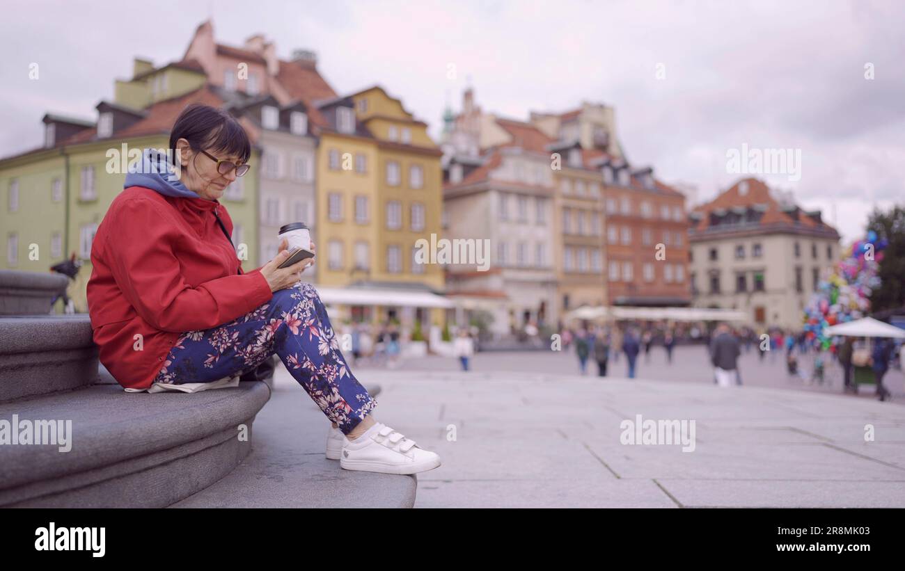 Elderly lady sits on the steps drinking coffee and using a smartphone in the historic center of an old European city. Palace Square, Warsaw Old Town Stock Photo