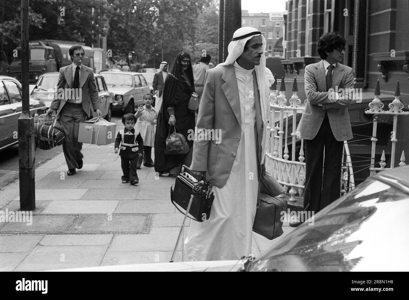 Arab Family in London with minder preparing to go home with stuff they have bought 1970s UK. Middle Eastern people came to Britain for subsidised health care in Harley Street clinics. They mainly stayed in cheap hotels in Earls Court. An Arab family leaving, the woman is wearing a Battoulah face mask indicated that she is married.  They are with their children. A British helper carries their bags, he's walking towards the car that takes them to the airport. Earls Court, London, England circa 1977 70s UK HOMER SYKES Stock Photo