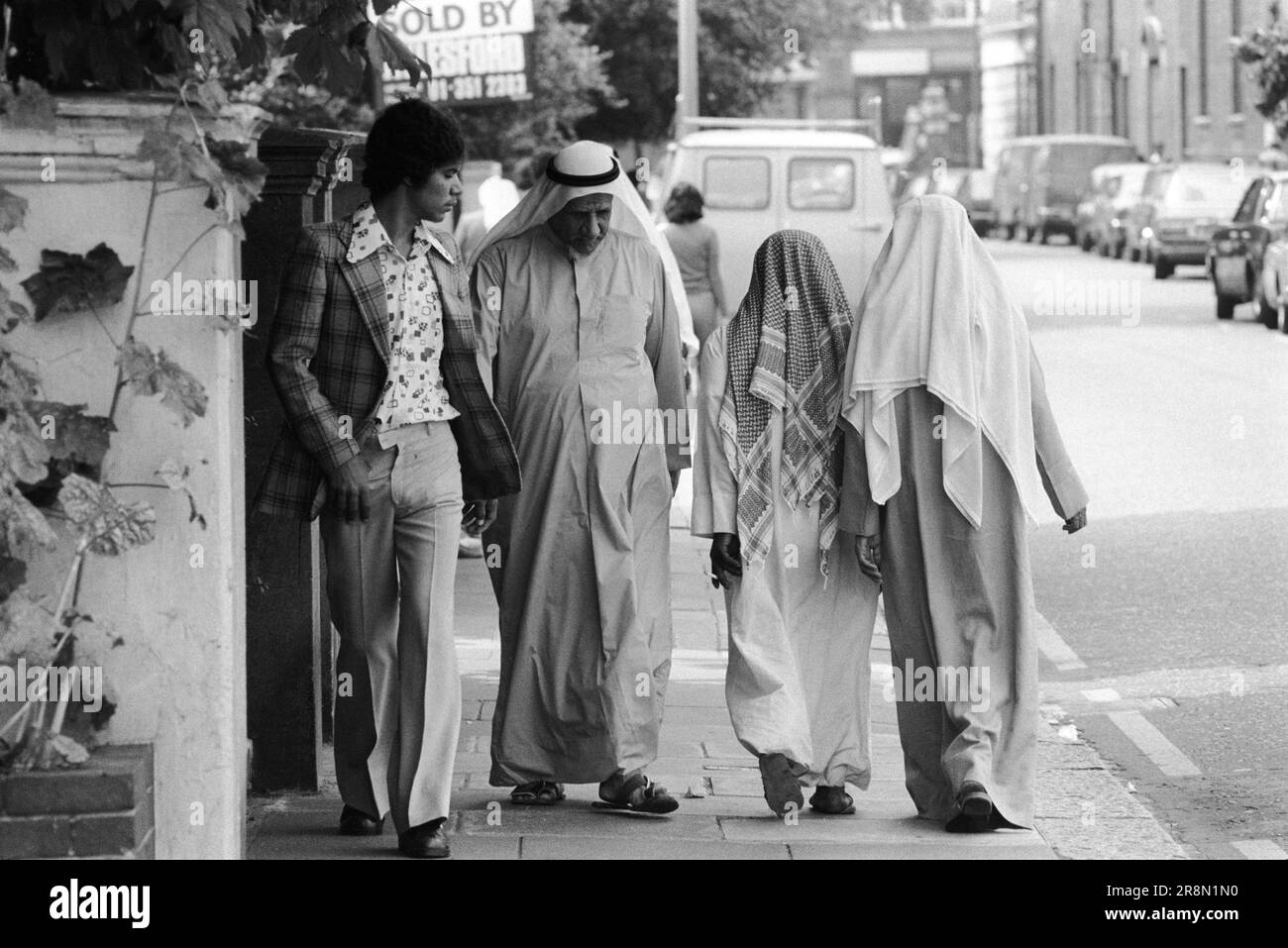 Poor Arab men walking around street of Earls Court London 1970s UK. Middle Eastern people came to Britain for subsidised health care in Harley Street clinics. They mainly stayed in the Earls Court area. Four men, a younger man in western dress and the other three men wearing the traditional white robes called a thoub, thobe, dishdasha or kandora and traditional Arab headdress called the kaffiyeh or ghutra. Earls Court, London, England circa 1977 70s UK HOMER SYKES Stock Photo