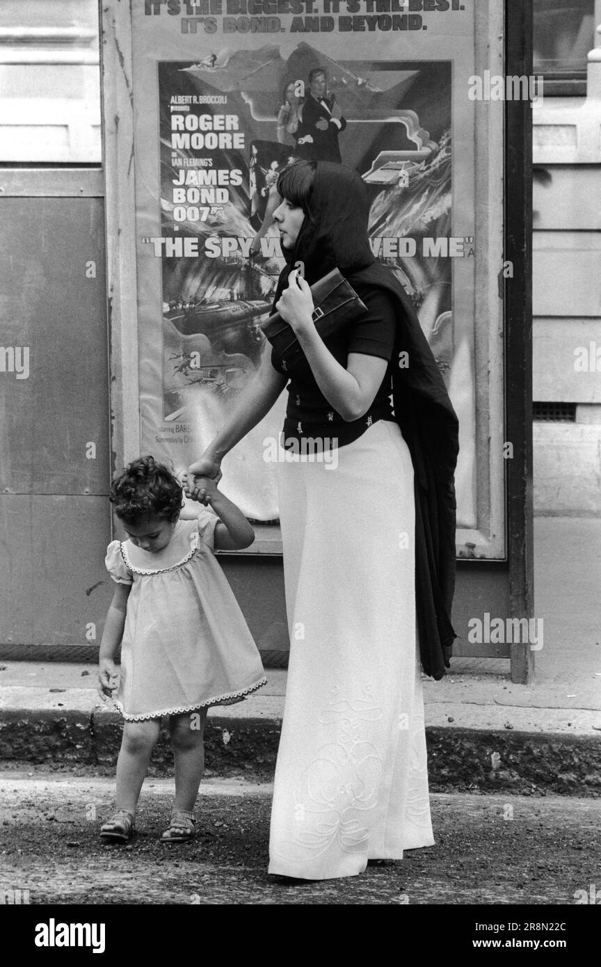 Muslim mother and child Poor Arabs in London 1970s. Middle Eastern people came to Britain for subsidised healthcare in Harley Street clinics. They mainly stayed in cheap hotels in Earls Court. Mother and daughter. The film poster is for The Spy Who Loved Me, staring Roger Moore as James Bond. Earls Court, London, England circa 1977 70s UK HOMER SYKES Stock Photo