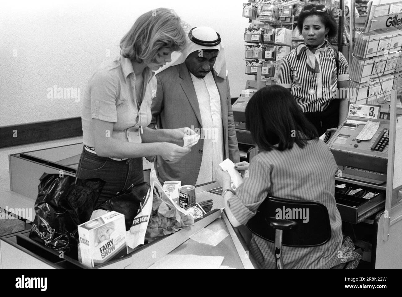 Poor Arabs in London 1970s Shopping. Middle Eastern people came to Britain for subsidised healthcare in Harley Street clinics. They mainly stayed in cheap hotels in Earls Court. Arab man Earls Court supermarket  two check out women help him with shopping and paying the bill. Earls Court, London, England circa 1977. 70s UK HOMER SYKES Stock Photo