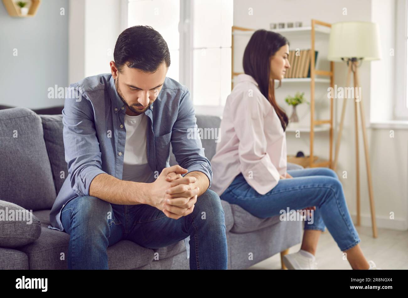 Upset young man sitting on sofa and a woman in background at home. Quarrel and divorce concept. Stock Photo