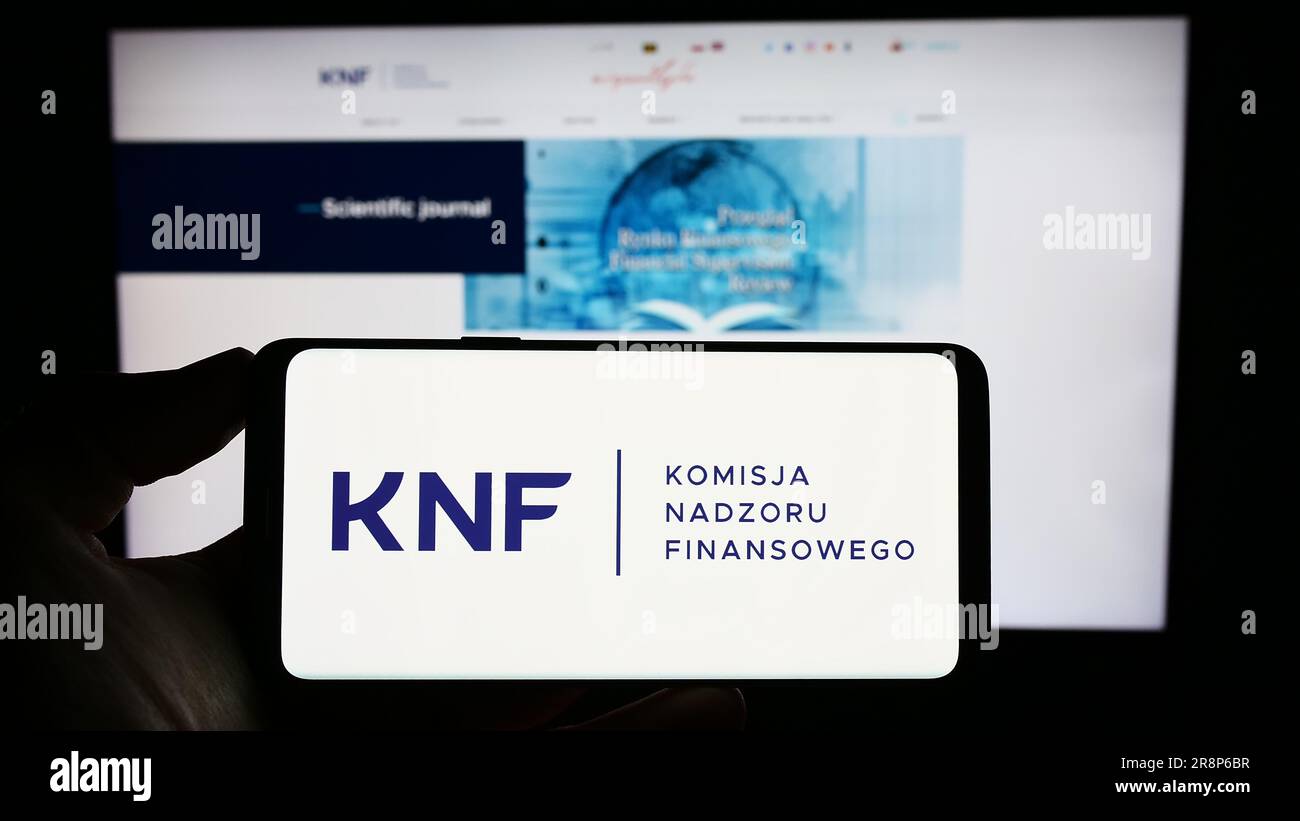 Person holding mobile phone with logo of authority Komisja Nadzoru Finansowego (KNF) on screen in front of web page. Focus on phone display. Stock Photo