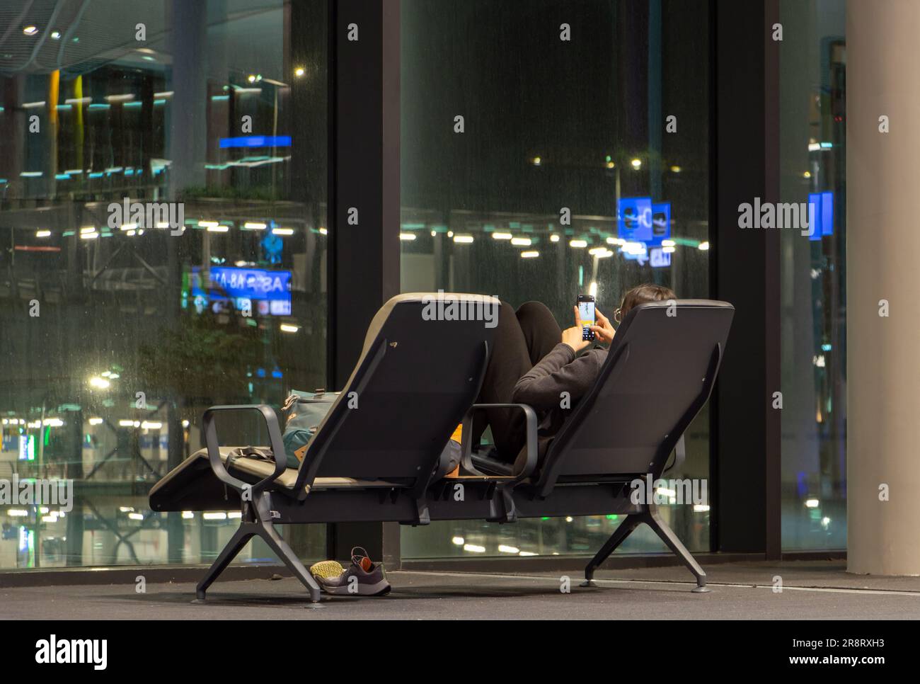 A young woman with a mobile phone is sitting opposite the window in the night hall, international airport. Stock Photo