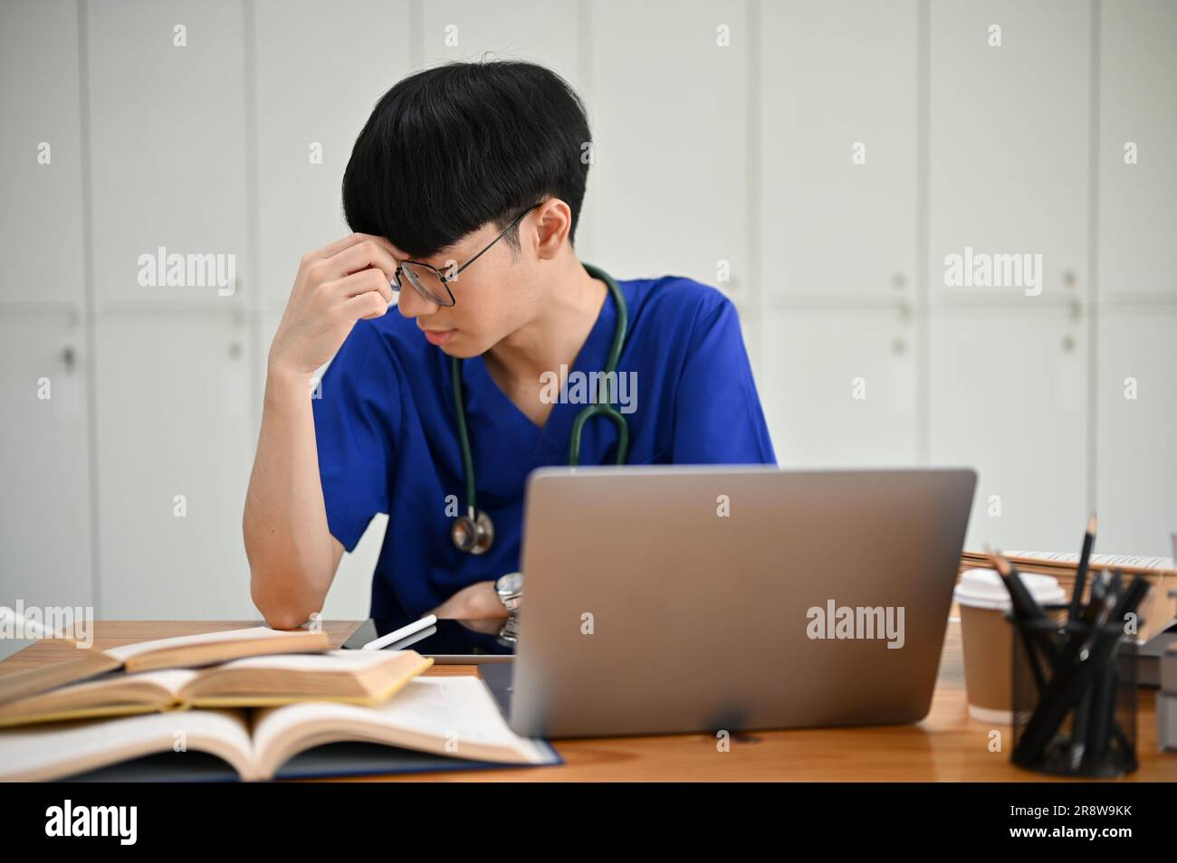 A tired young Asian male medical student in scrubs suffers from ...