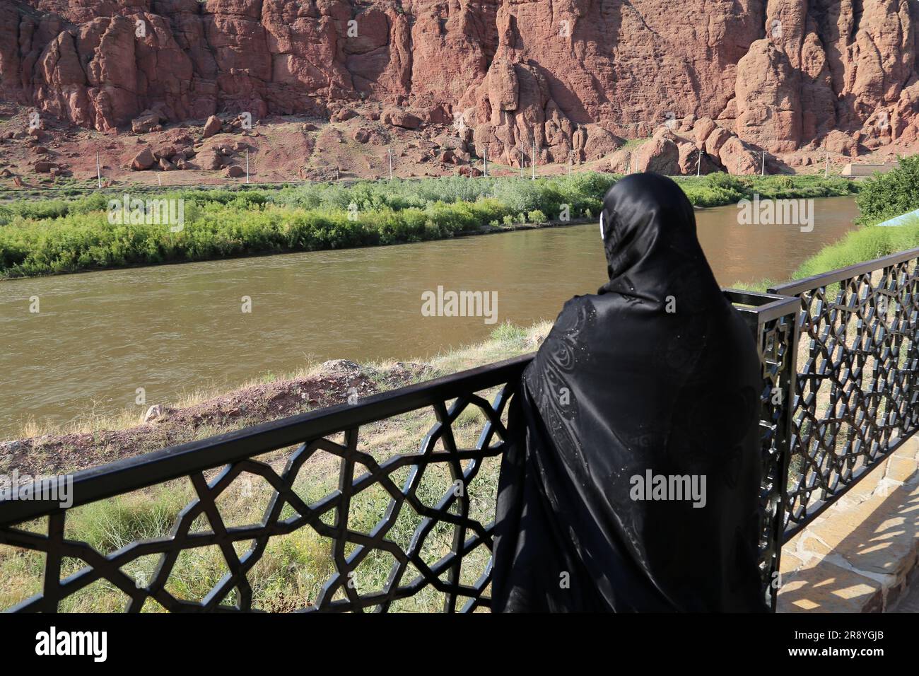 June 21, 2023, Jolfa, East Azerbaijan, Iran: A veiled woman in a black chador looks at the Aras River at the border between northwestern Iran and Azerbaijan. The Aras (the Araks, Arax, Araxes, or Araz) is a river in the Caucasus. It rises in eastern Turkey and flows along the borders between Turkey and Armenia, between Turkey and the Nakhchivan exclave of Azerbaijan, between Iran and both Azerbaijan and Armenia, and, finally, through Azerbaijan where it flows into the Kura River. It drains the south side of the Lesser Caucasus Mountains while the Kura drains the north side of the Lesser Caucas Stock Photo