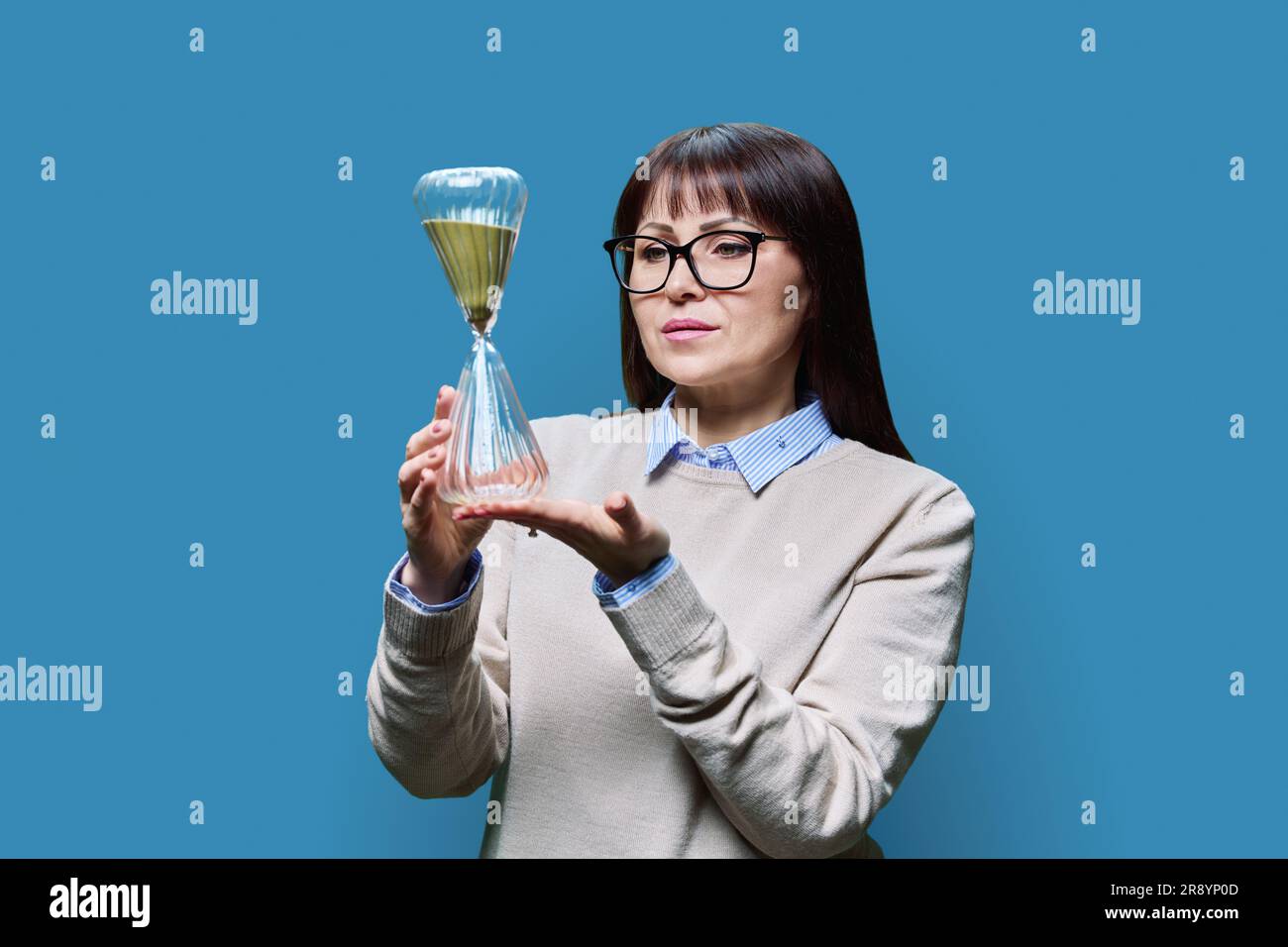 Middle aged woman with hourglass on blue studio background Stock Photo