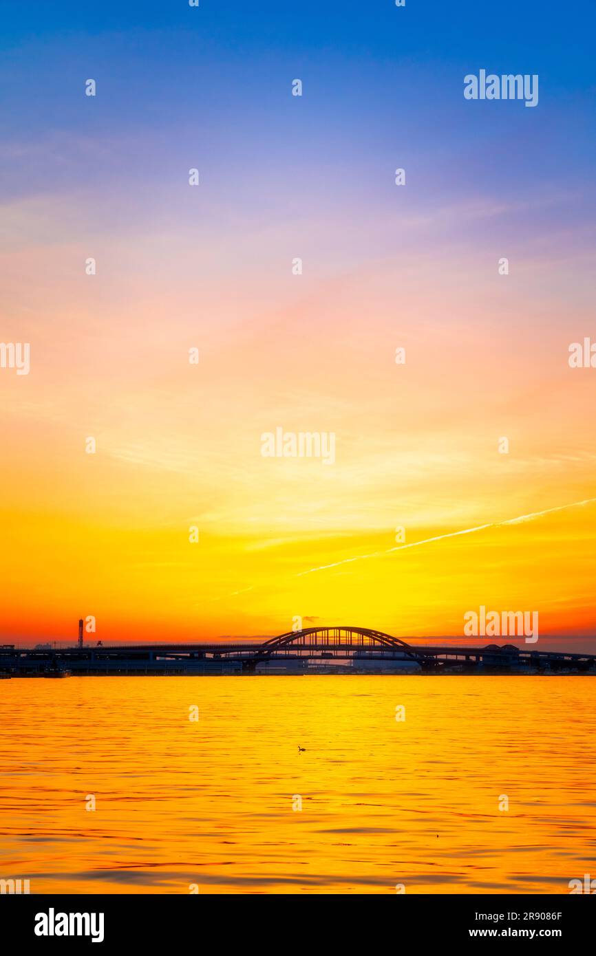 A vivid orange dawn sky behind the silhouette of the doubledeck arched Kobe Bridge connecting