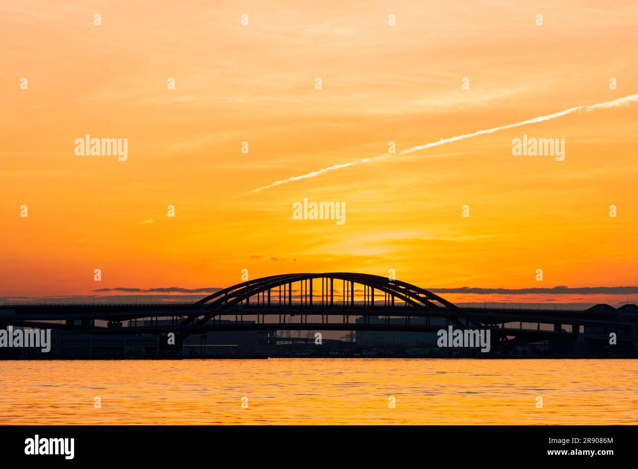 A vivid orange dawn sky behind the silhouette of the double-deck arched Kobe Bridge connecting the main land with Port Island at Kobe in Japan. Stock Photo