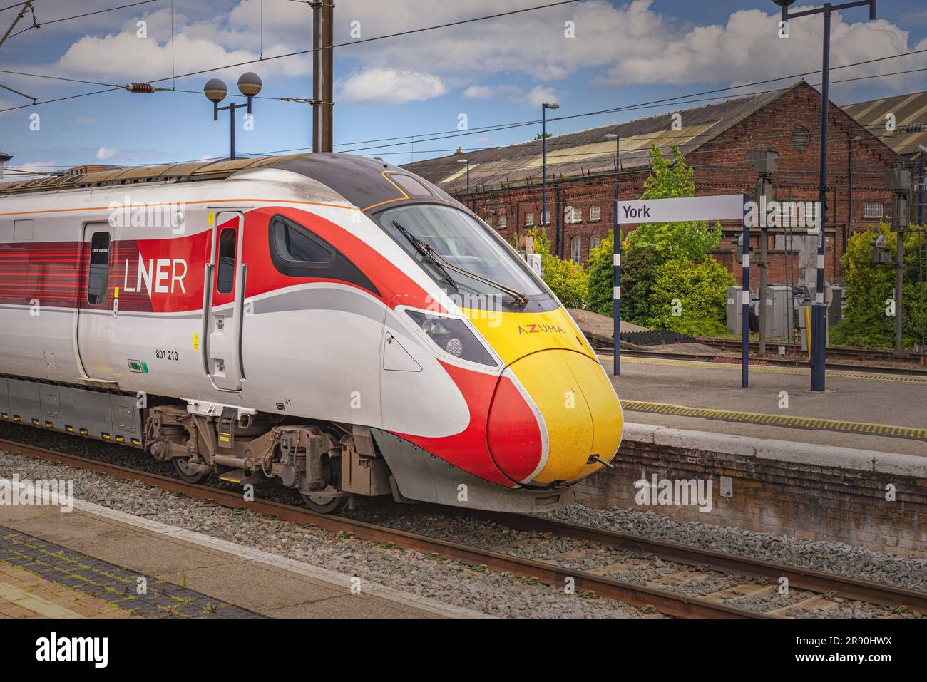 A train stands at a railway station platform beside a location sign. A