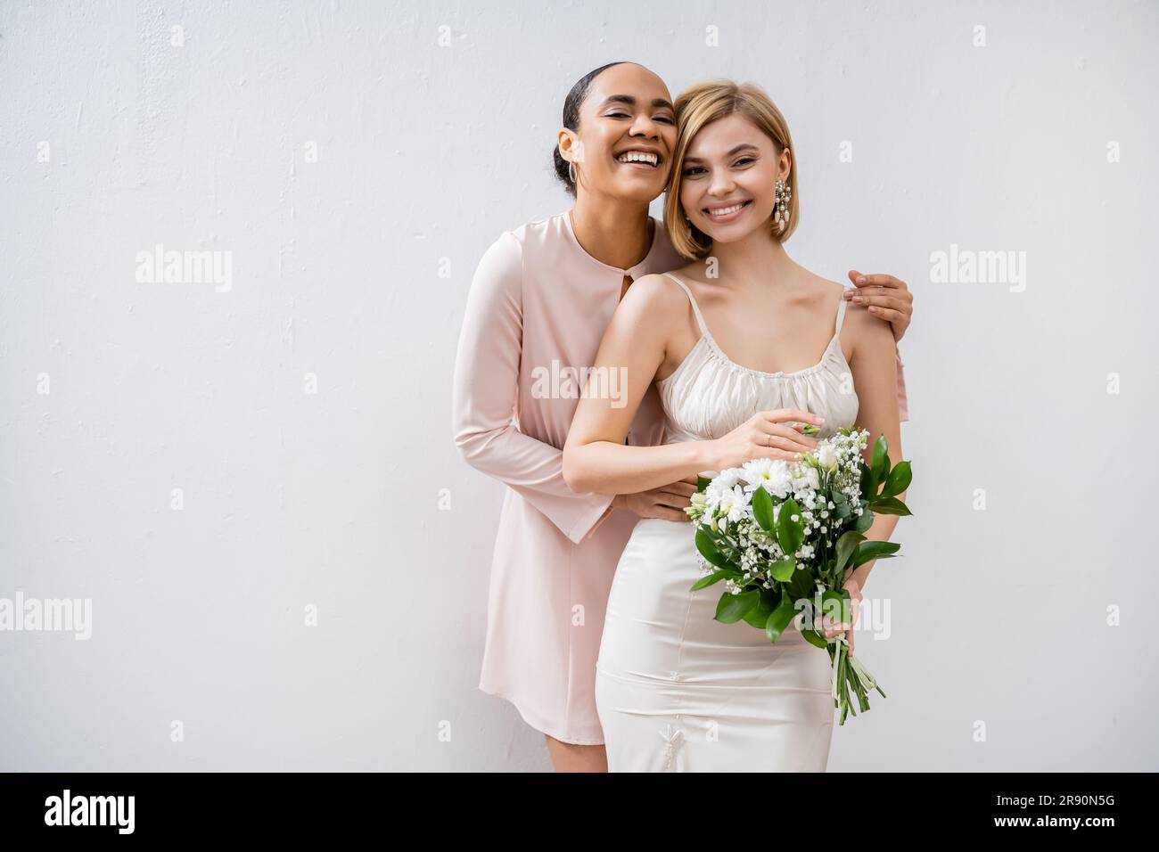 bridal shower, happy bride with bridesmaid, cheerful interracial women, wedding dress and bridesmaid gown, positive african american woman hugging eng Stock Photo