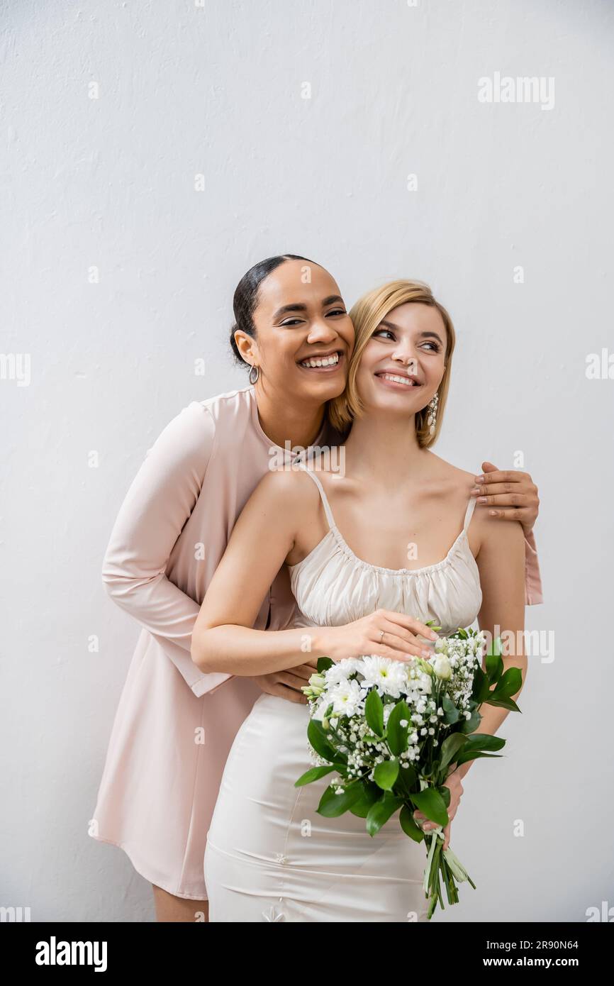bridal party, happy bride with bridesmaid, cheerful interracial women, wedding dress and bridesmaid gown, african american woman hugging engaged frien Stock Photo