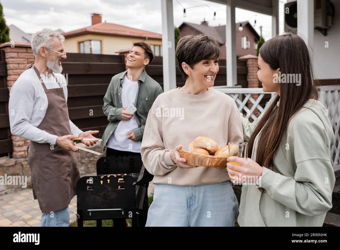 celebration of parents day, middle aged mother holding buns and talking with teenage daughter, father and son preparing food on barbecue grill, laught Stock Photo