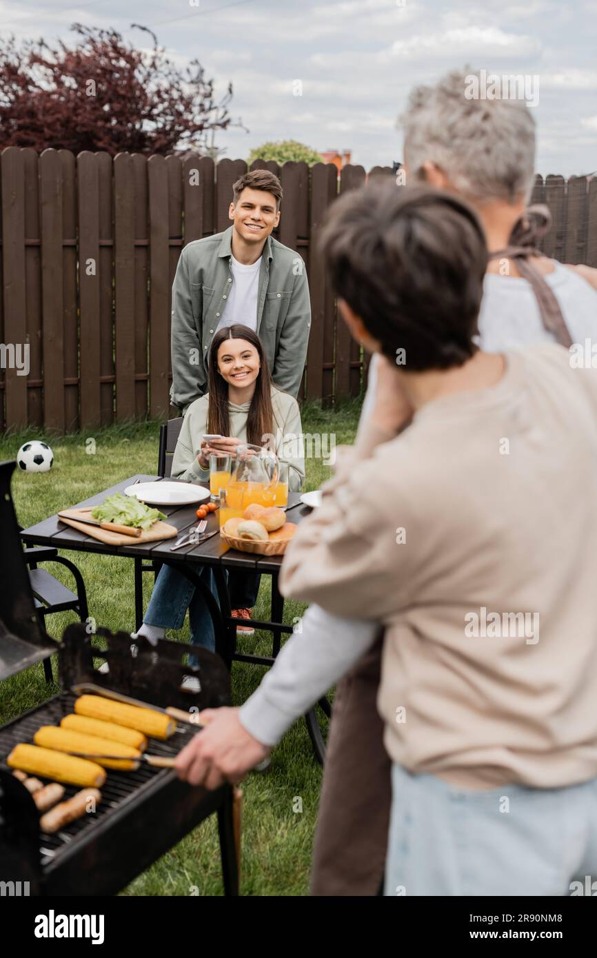 parents day celebration, happy siblings looking at father and mother on blurred background, family bbq, grill party, using smartphone, preparing food Stock Photo
