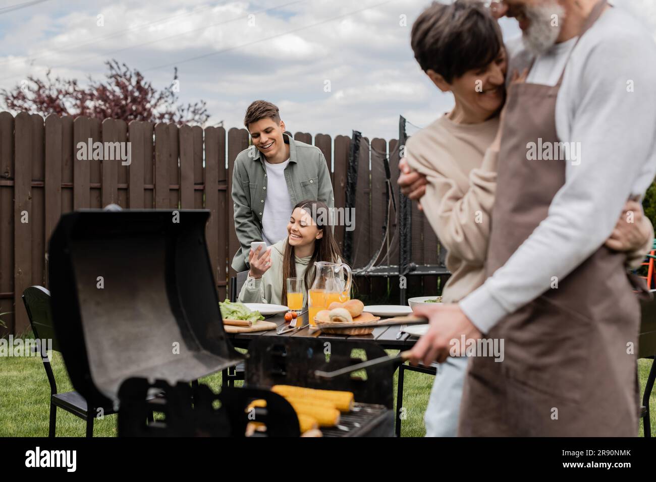 happy teenage girl showing something on smartphone to young adult brother, digital age, father preparing food on bbq grill, barbecue party, parents da Stock Photo