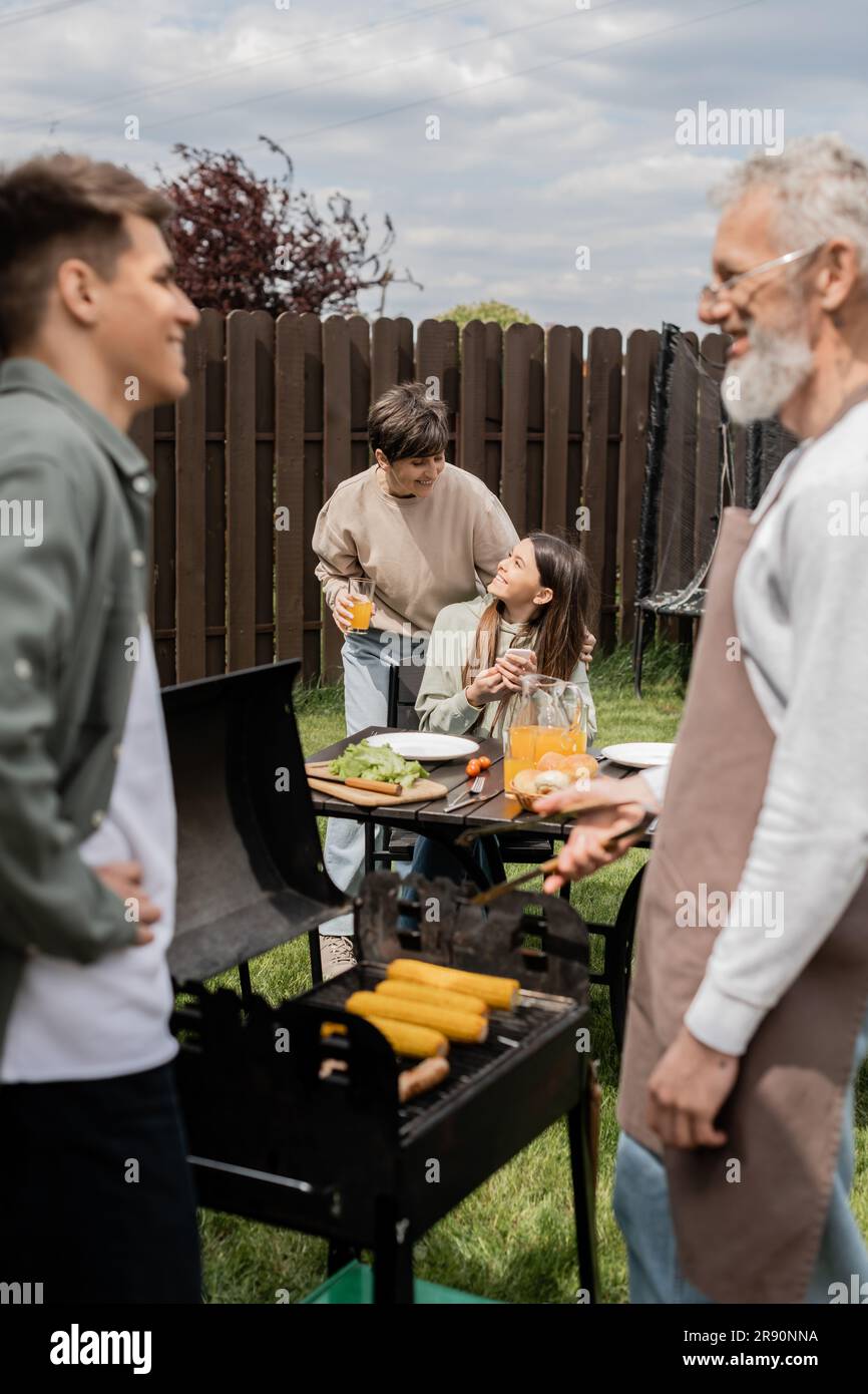 cheerful teenage girl holding smartphone and looking at mother digital age, father and son preparing food on bbq grill, barbecue party, parents day ce Stock Photo