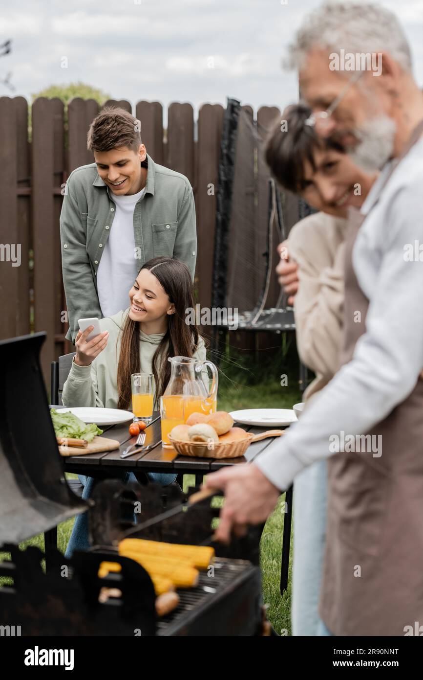 happy teenage girl showing something on smartphone to young adult brother, digital age, father preparing food on bbq grill, barbecue party, parents da Stock Photo