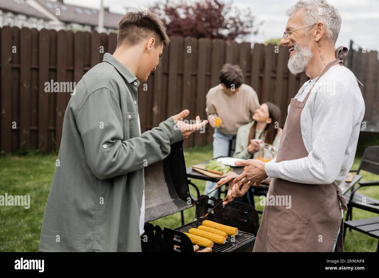 tattooed and bearded father and son preparing food on bbq grill, grilling corn, communication, family bbq party, middle aged, happy parents day, trans Stock Photo