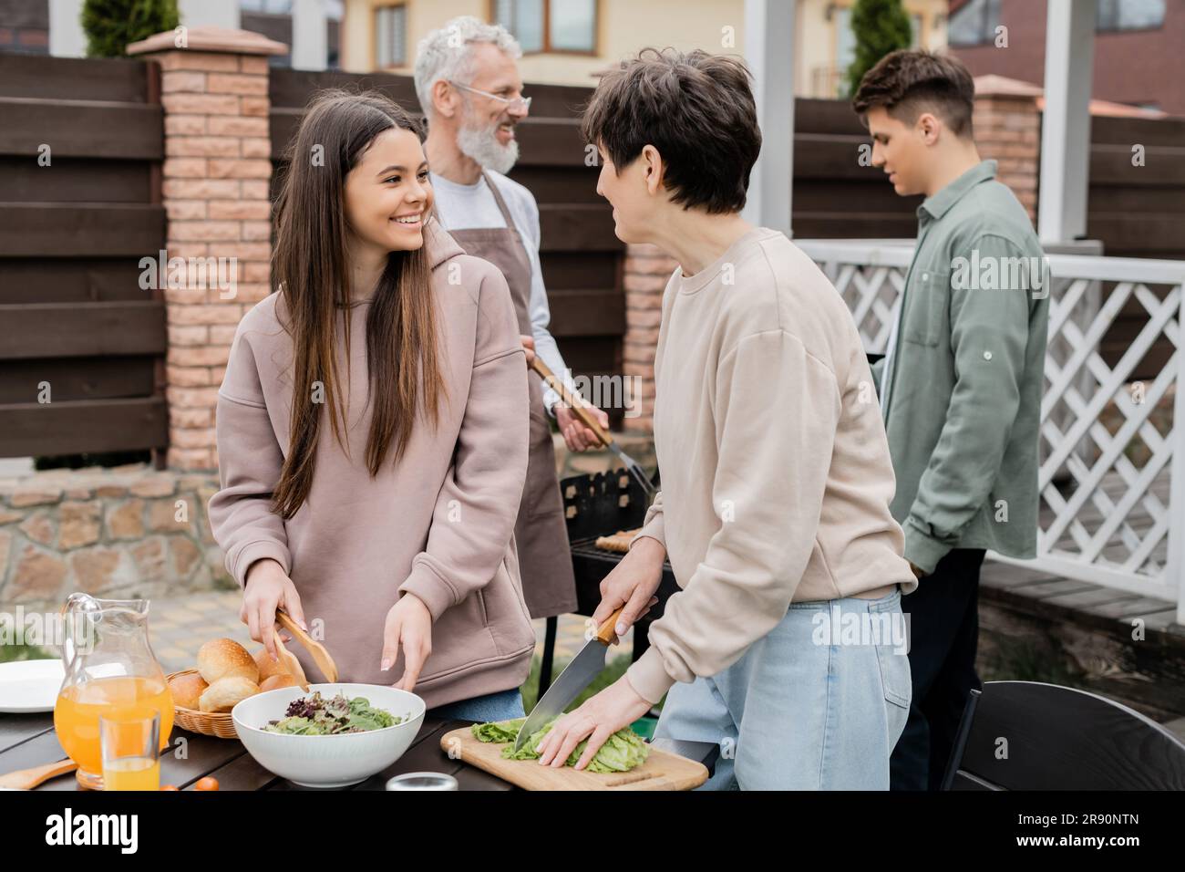 cheerful mother and teenage daughter making salad, father and son preparing food on bbq grill, family house backyard, suburban life, happy parents day Stock Photo