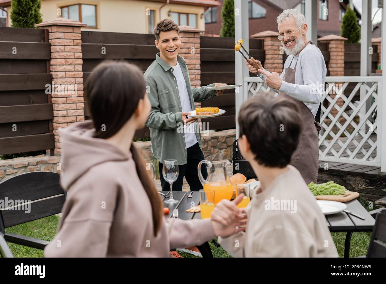 bearded dad and his young adult son preparing food on bbq grill, family bbq party, grilled corn, middle aged man looking at wife and teenage daughter Stock Photo