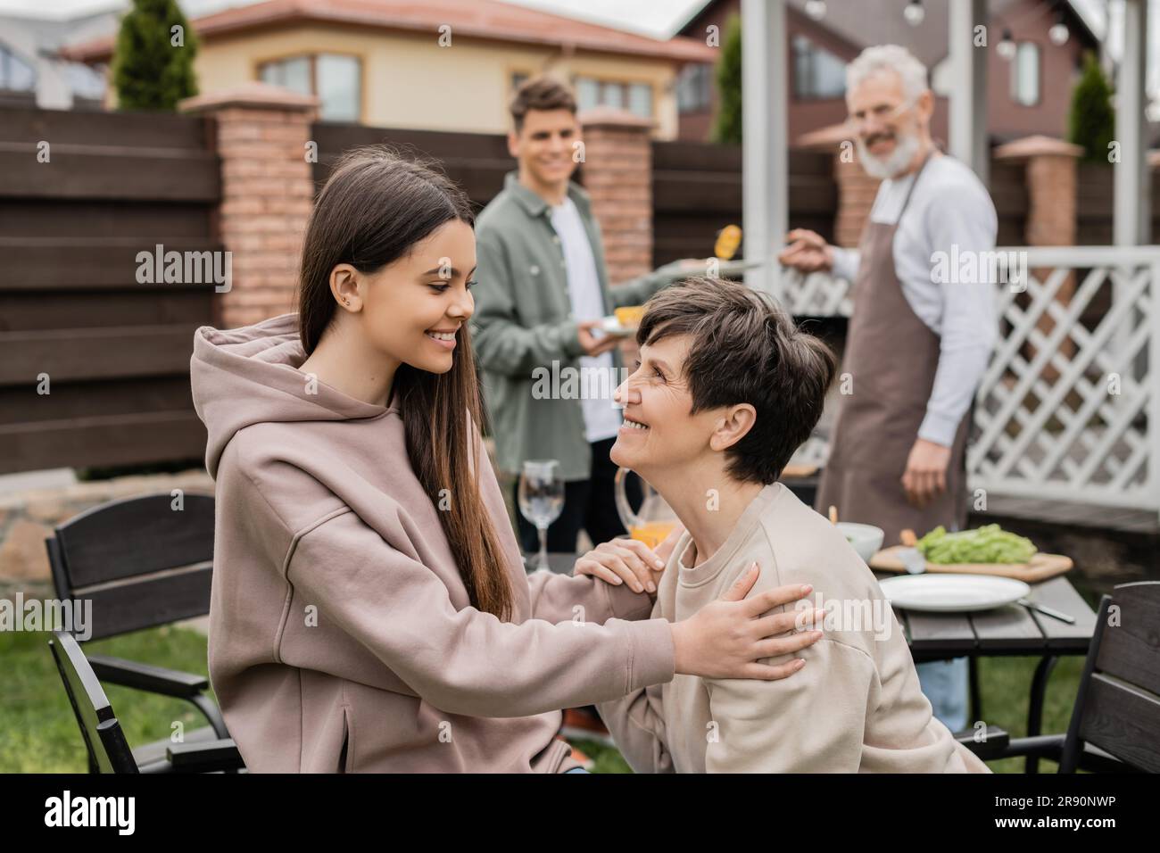 cheerful teenage girl talking to middle aged mother during family grill party, bonding, father and son preparing food on bbq grill, celebrating parent Stock Photo