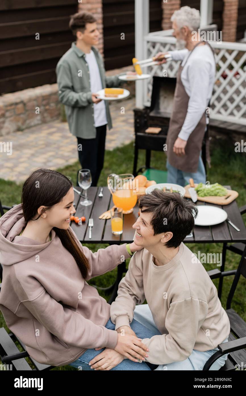 joyful teenage girl talking to middle aged mother during family grill party, summer food, father and son preparing food on bbq grill, celebration of p Stock Photo