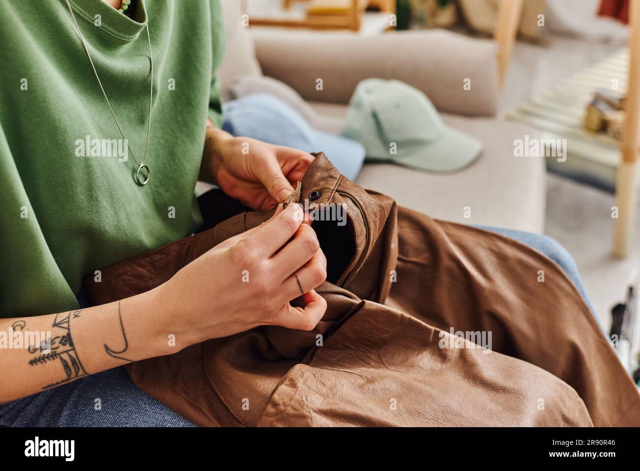 cropped view of young tattooed woman buttoning leather pants while sitting on couch near wardrobe items during decluttering process, sustainable livin Stock Photo