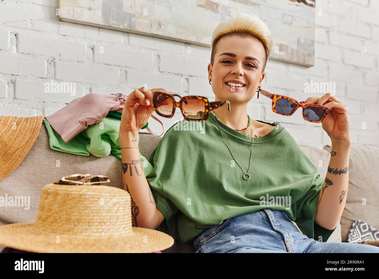 happy young woman sitting on couch near straw hat and clothing while showing stylish sunglasses, trendy hairstyle, tattoo, sustainable living and mind Stock Photo