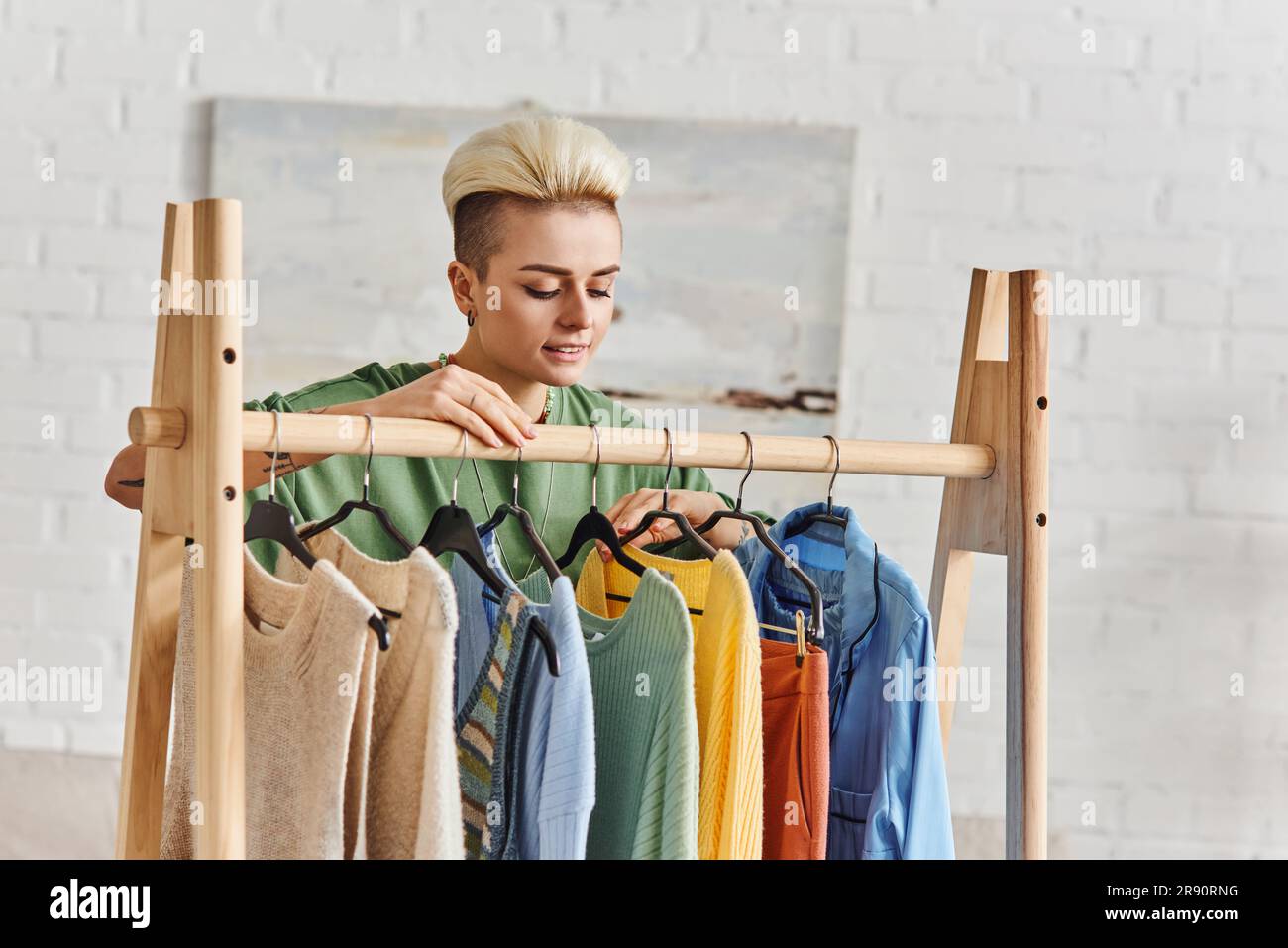 young and happy woman looking at fashionable casual clothes on rack in living room, thrift store finds, second-hand, conscious lifestyle, sustainable Stock Photo