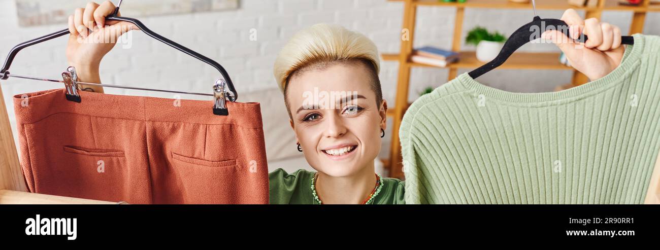 pleased young woman with radiant smile and trendy hairstyle holding hangers with pants and jumper while looking at camera at home, sustainable fashion Stock Photo