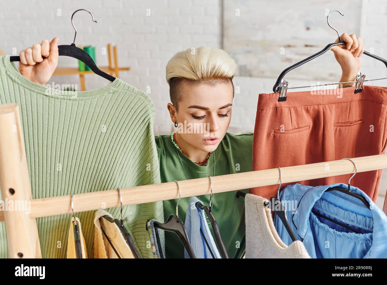 thoughtful woman with trendy hairstyle holding hangers with pants and jumper near rack with casual clothes, home decluttering, sustainable fashion and Stock Photo