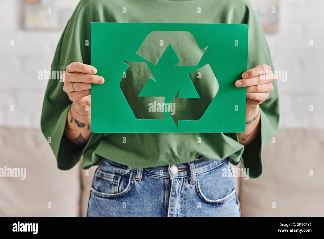 green living, partial view of young and tattooed woman in casual clothes holding green paper card with cutout recycled sign at home, sustainable and e Stock Photo