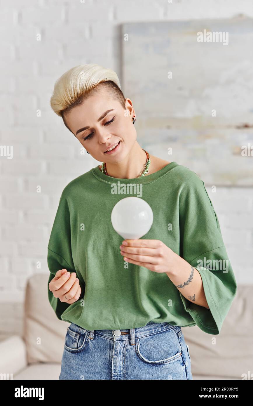 green living, young tattooed woman with trendy hairstyle and happy face holding energy saving light bulb at home, sustainable lifestyle and environmen Stock Photo