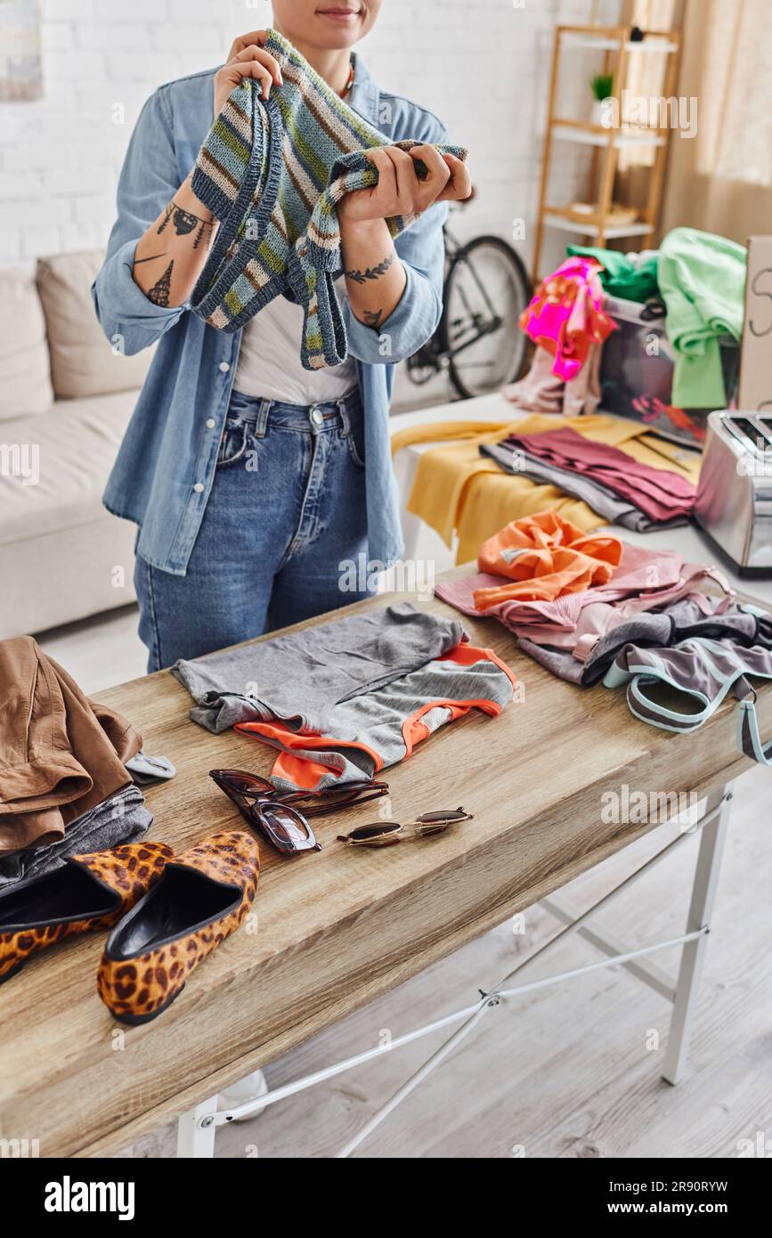 eco-friendly swaps, partial view of tattooed woman with knitted tank top near animal print shoes, sunglasses and casual clothes on table at home, sust Stock Photo