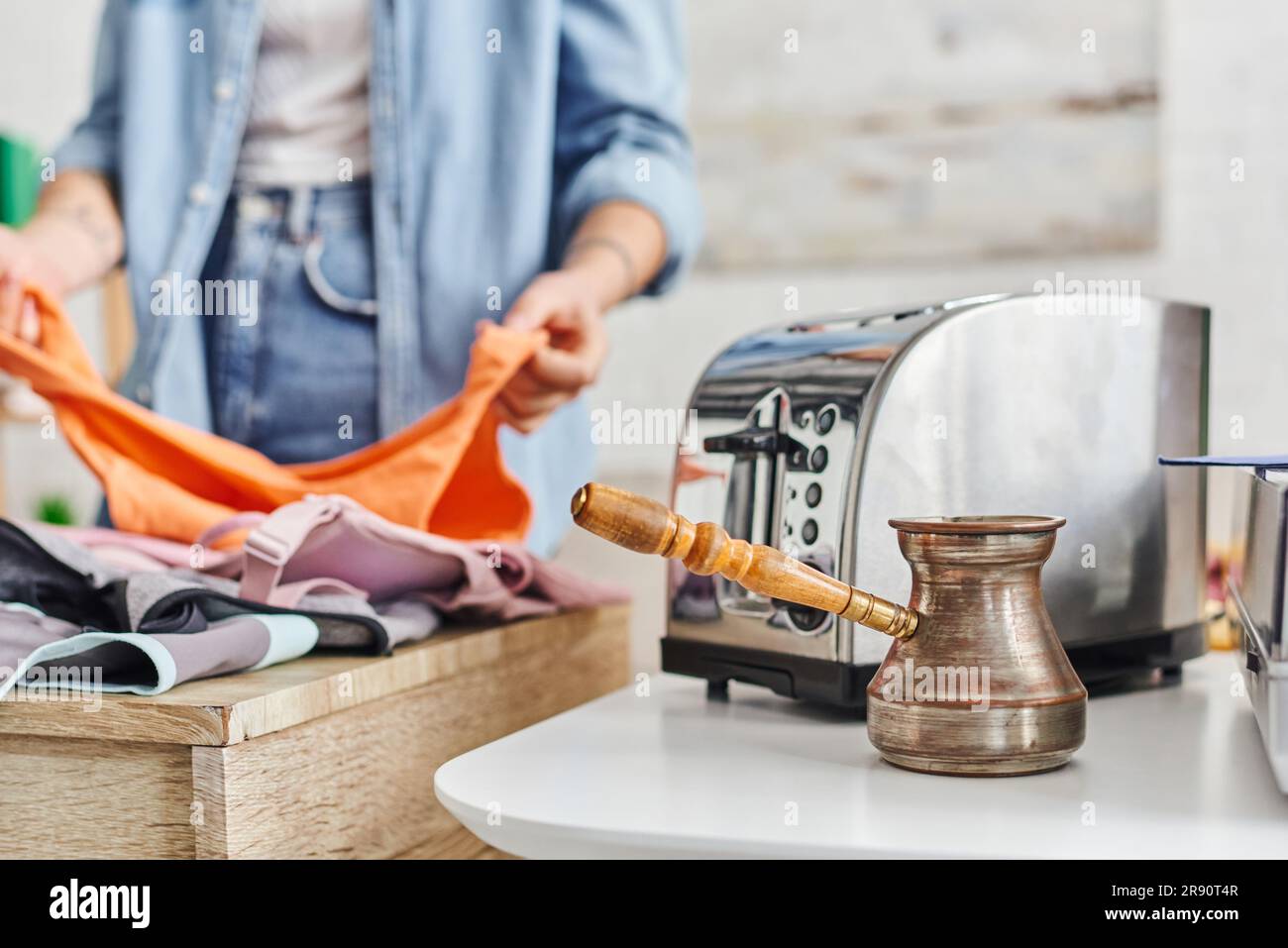 partial view of young woman sorting clothes near electric toaster and cezve, eco-friendly swaps, exchange market, blurred background, sustainable livi Stock Photo