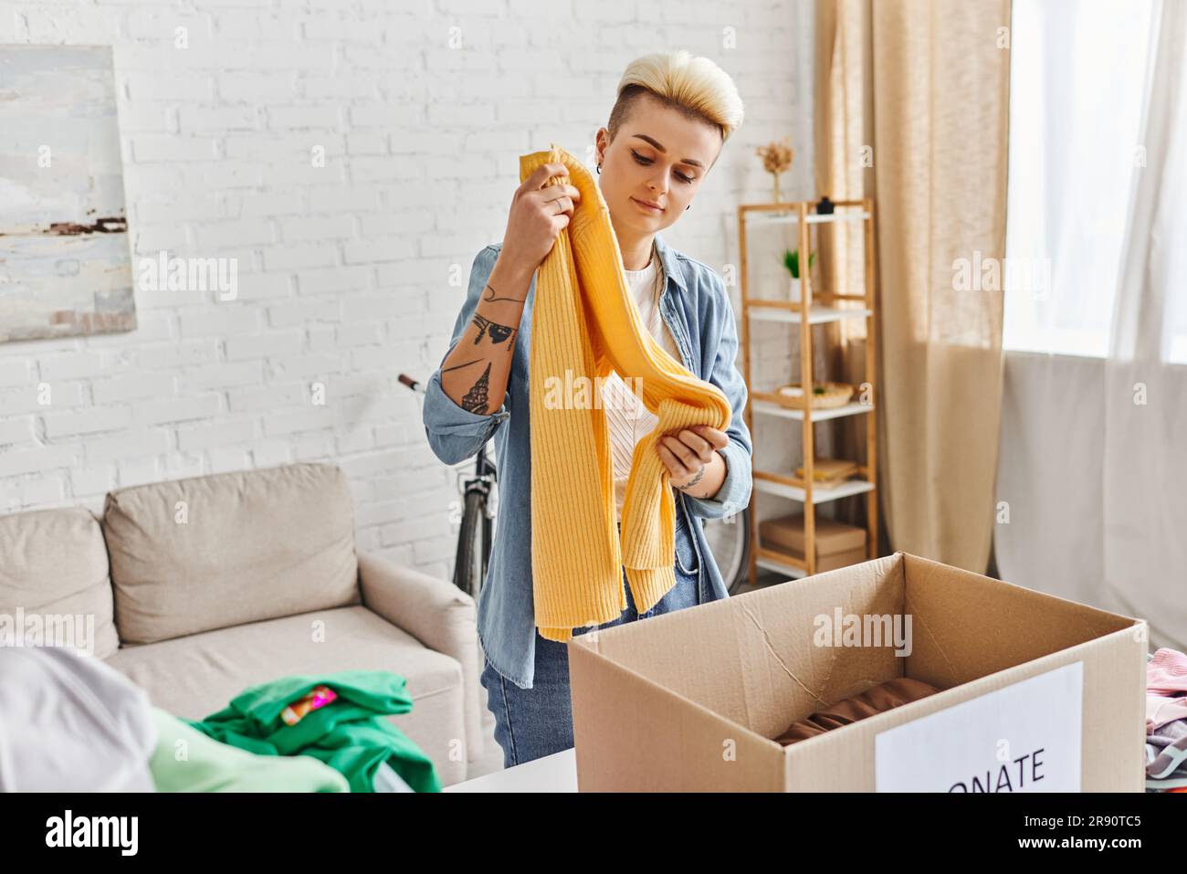 volunteering and charity, trendy and tattooed woman standing with yellow jumper near carton box while sorting clothing for donation at home, sustainab Stock Photo
