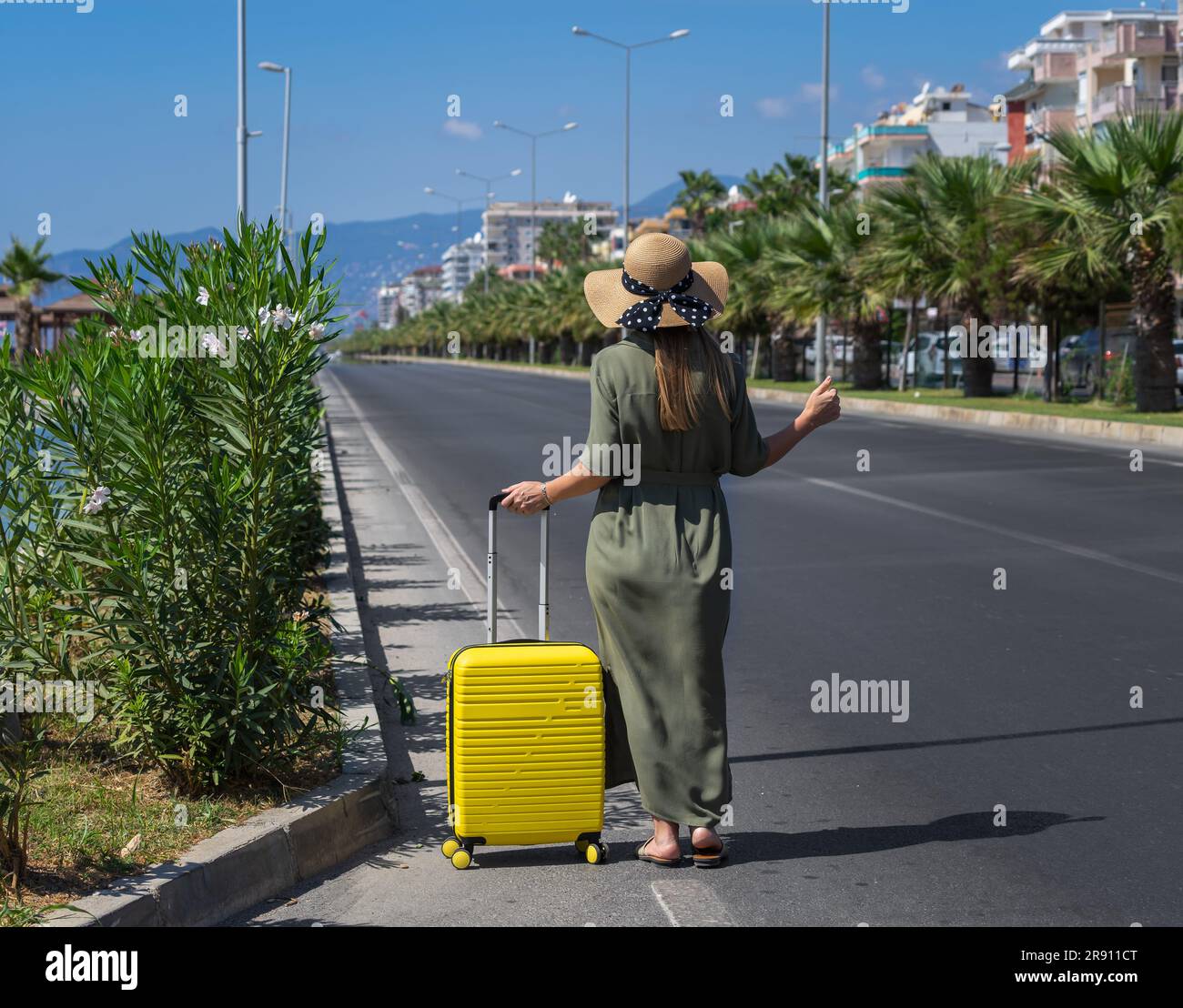 A girl with a yellow travel bag stands at the edge of the roadway. Stock Photo