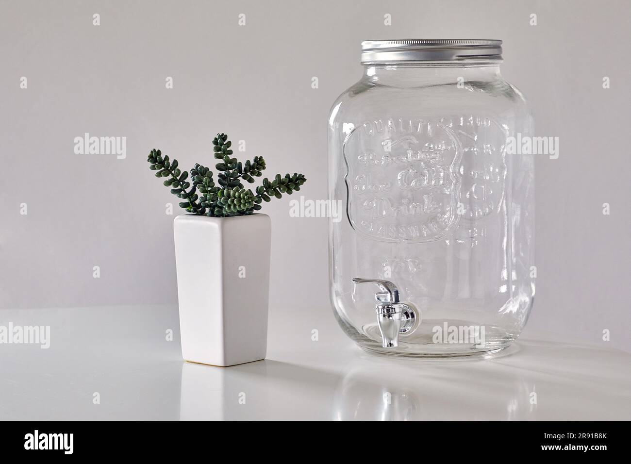 https://c8.alamy.com/comp/2R91B8K/white-table-with-succulent-plant-in-a-white-pot-and-empty-glass-water-dispenser-against-a-white-wall-2R91B8K.jpg