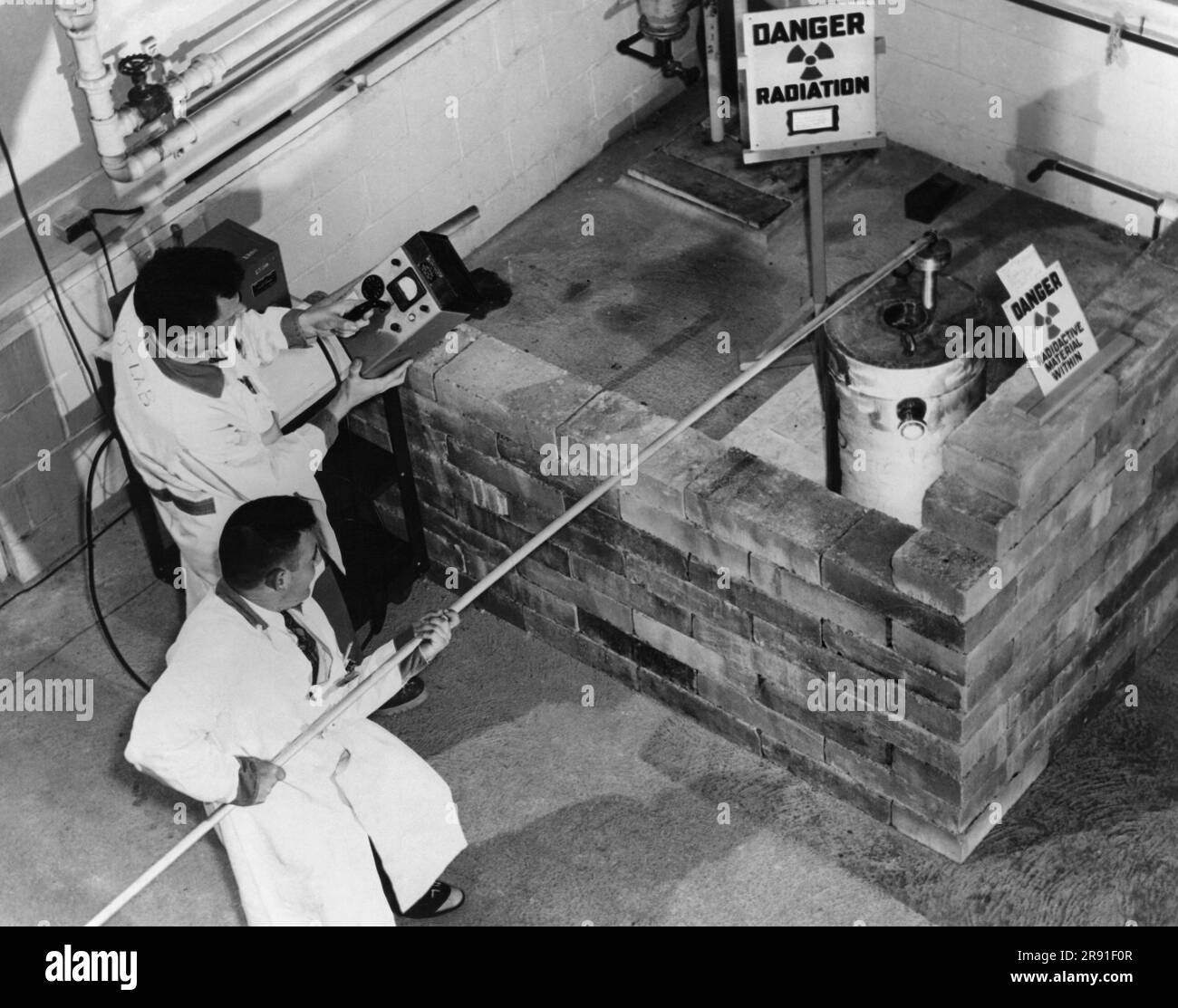 Upton, New York,  March, 1952 A technician at Brookhaven National Laboratory on Long Island stands behind a brick wall as he uses a long steel pole to remove the plug from a one and one-half ton lead shield containing radioactive tantalum while a health physicist uses a survey meter containing a Geiger Counter to make sure personnel are not exposed to dangerous amounts of radiation. Stock Photo