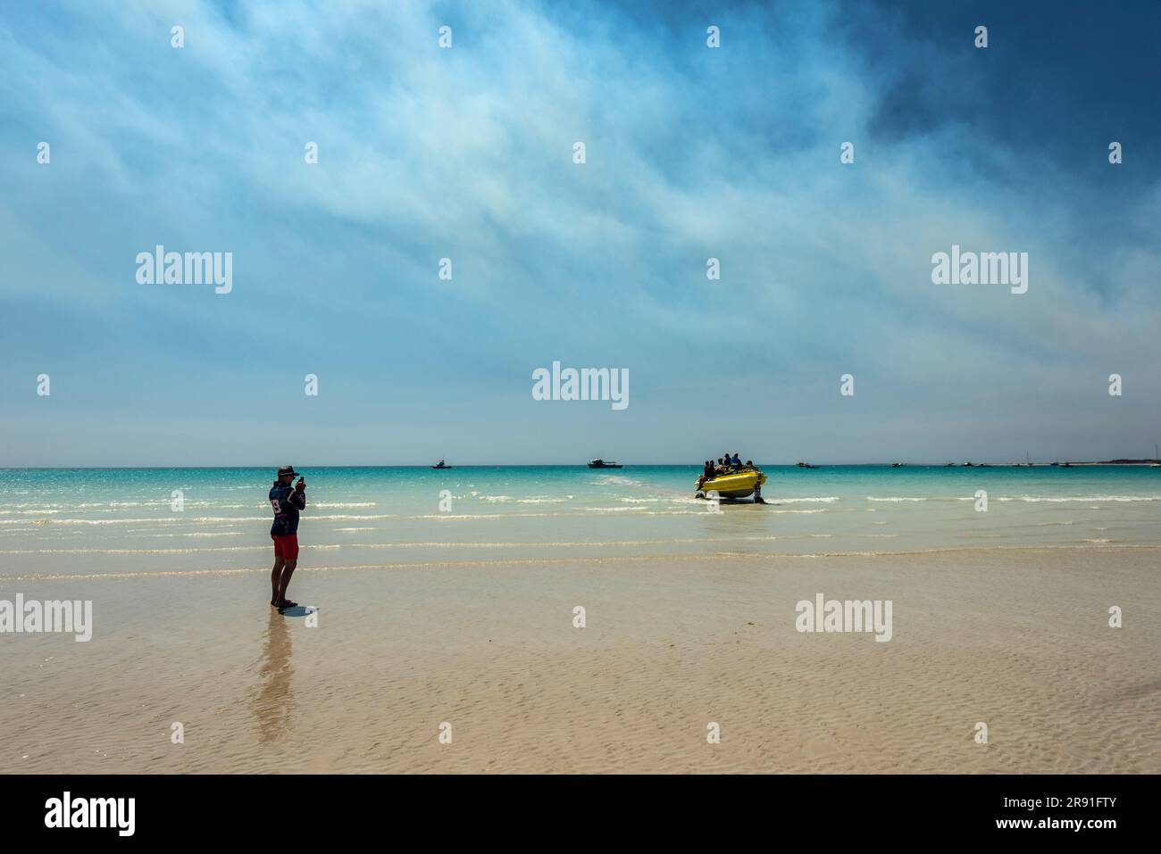 A person takes a photo of a boat on wheels emerging from the water on Cable Beach in Broome Western Australia Stock Photo