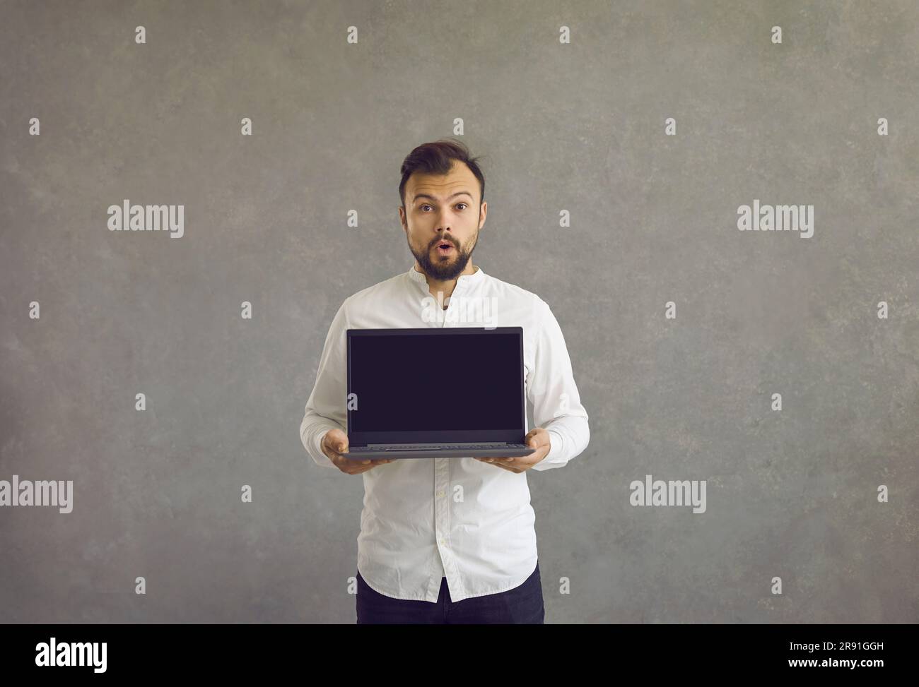 A young man stands with his mouth wide open, surprised, holding a laptop in his hands with a black screen. Stock Photo