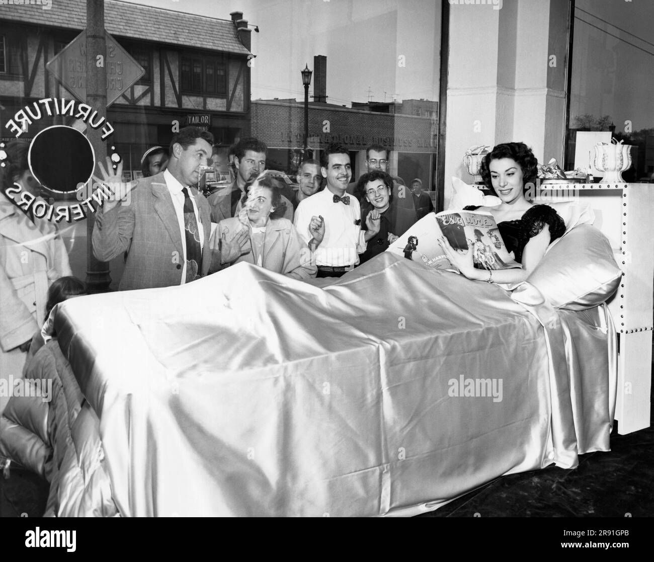 Chicago, Illinois:  October 10, 1951 A model reads an Esquire magazine in bed in the store window of Kay's Interiors to call attention to a new line of linens introduced by the store. She seems unperturbed by the rapt audience outside her window. Stock Photo