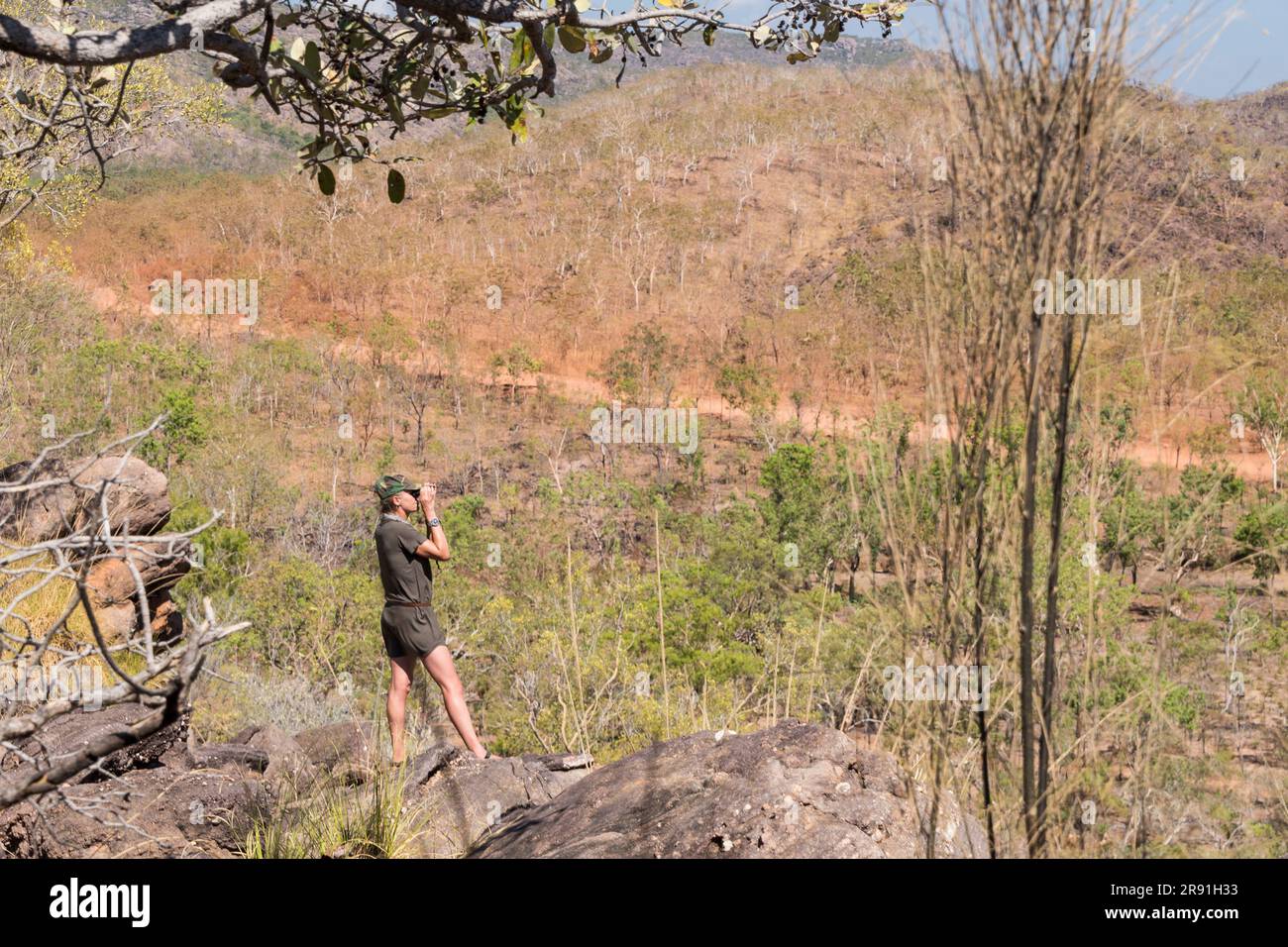 A woman in a sun hat looks through binoculars from a walking trail in the outback in Australia Stock Photo
