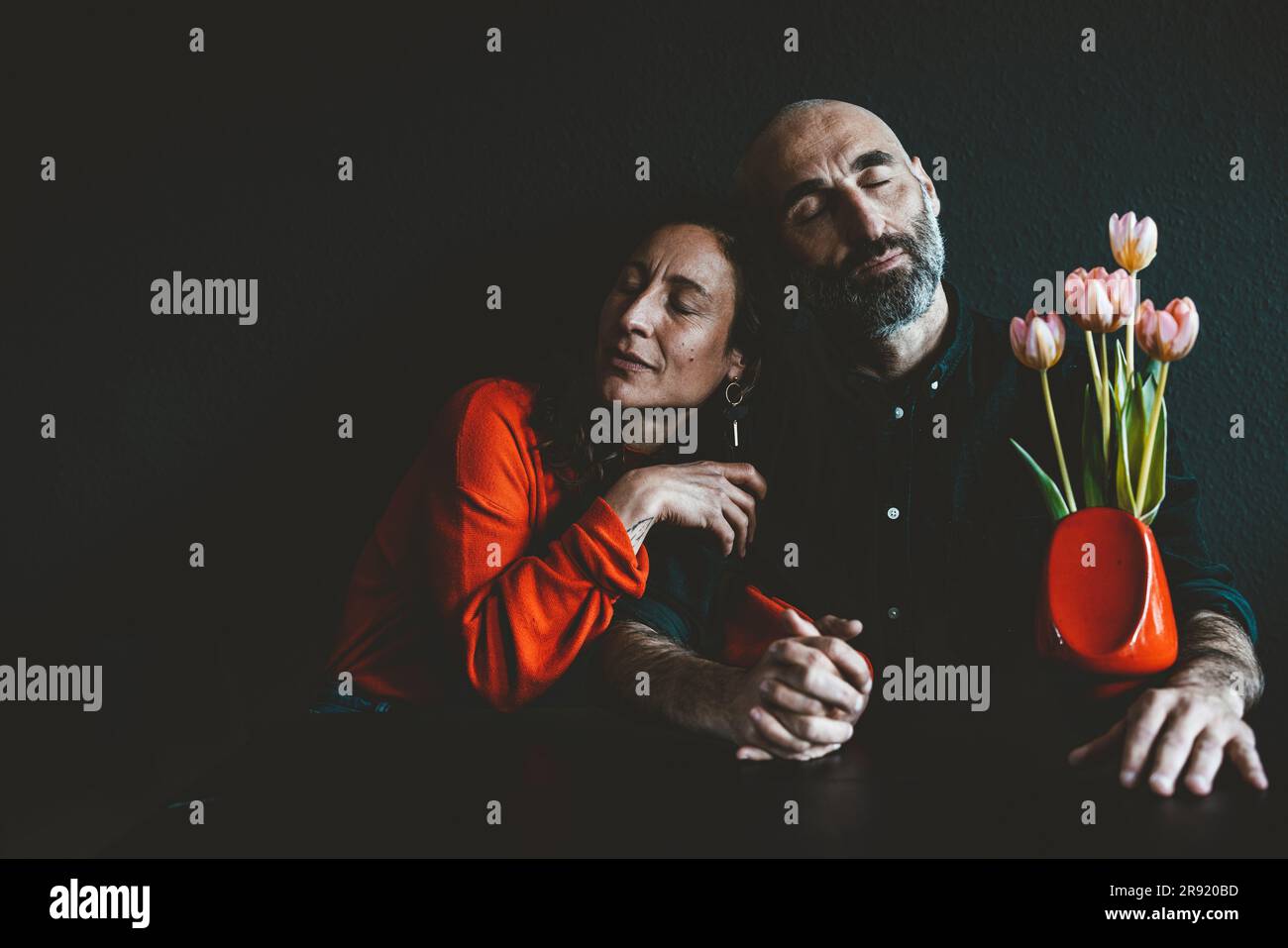 Couple with eyes closed sitting with red flower vase together in front