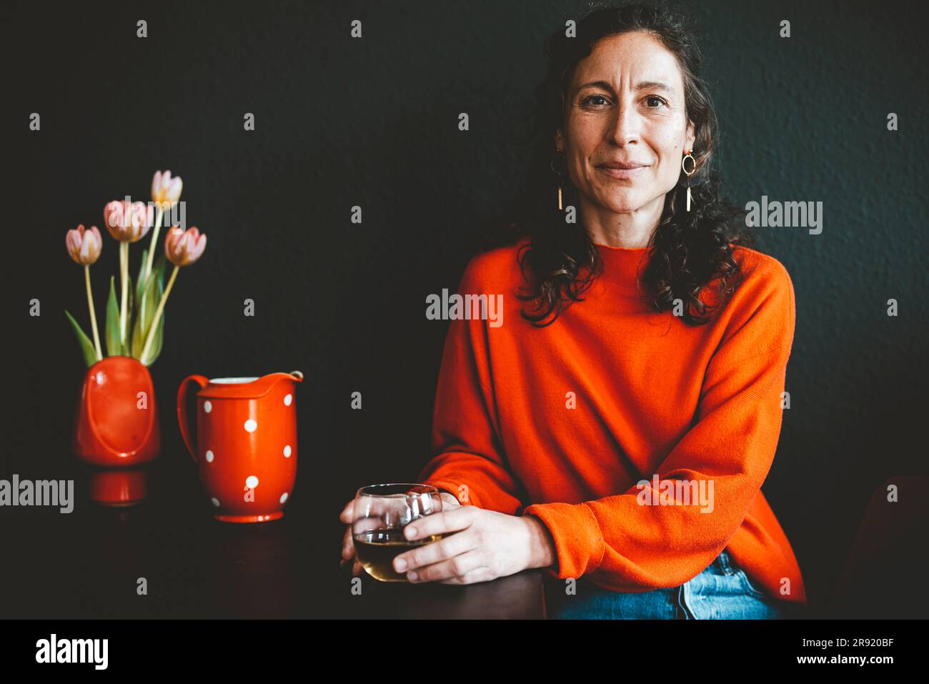 Smiling woman with drink sitting at table with red flower vase in front of black wall Stock Photo