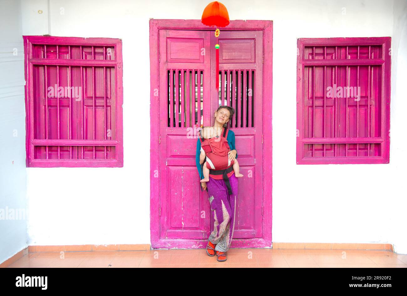 Mature woman poses in front of a pink door with baby being carried in Penang Malaysia Stock Photo