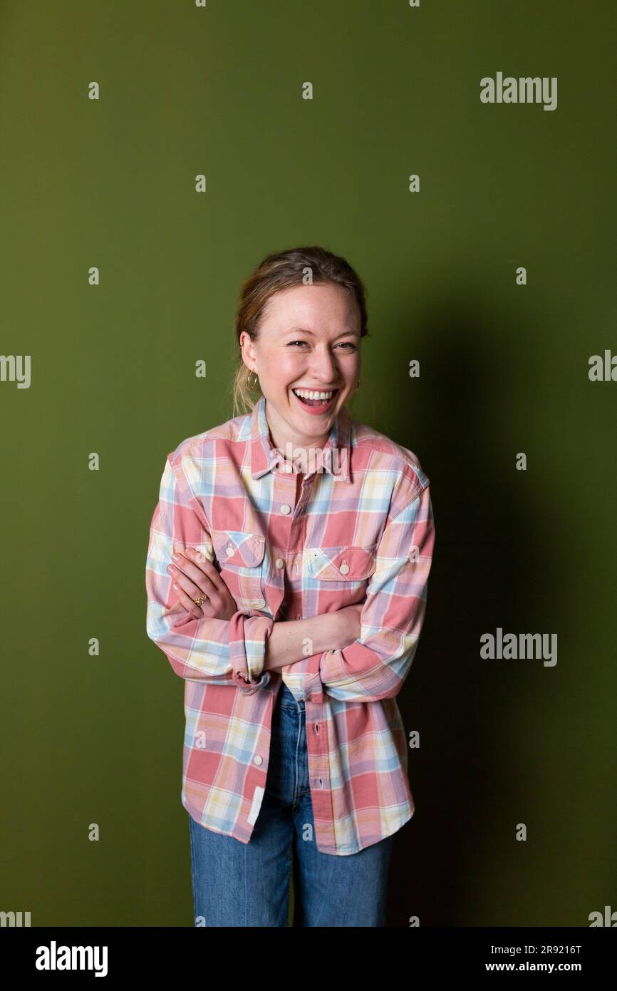 Young woman in plaid shirt laughing with arms crossed in front of green wall Stock Photo